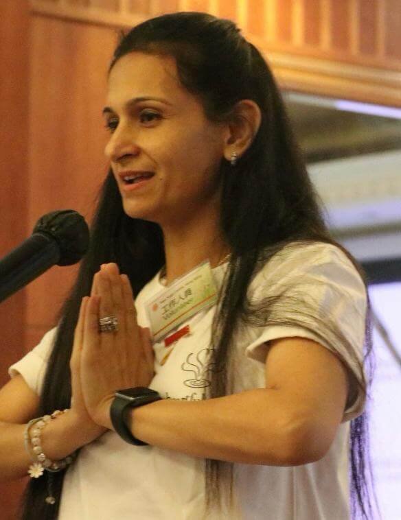 Neha Khera Saluja speaking at a yoga event with hands together in greeting